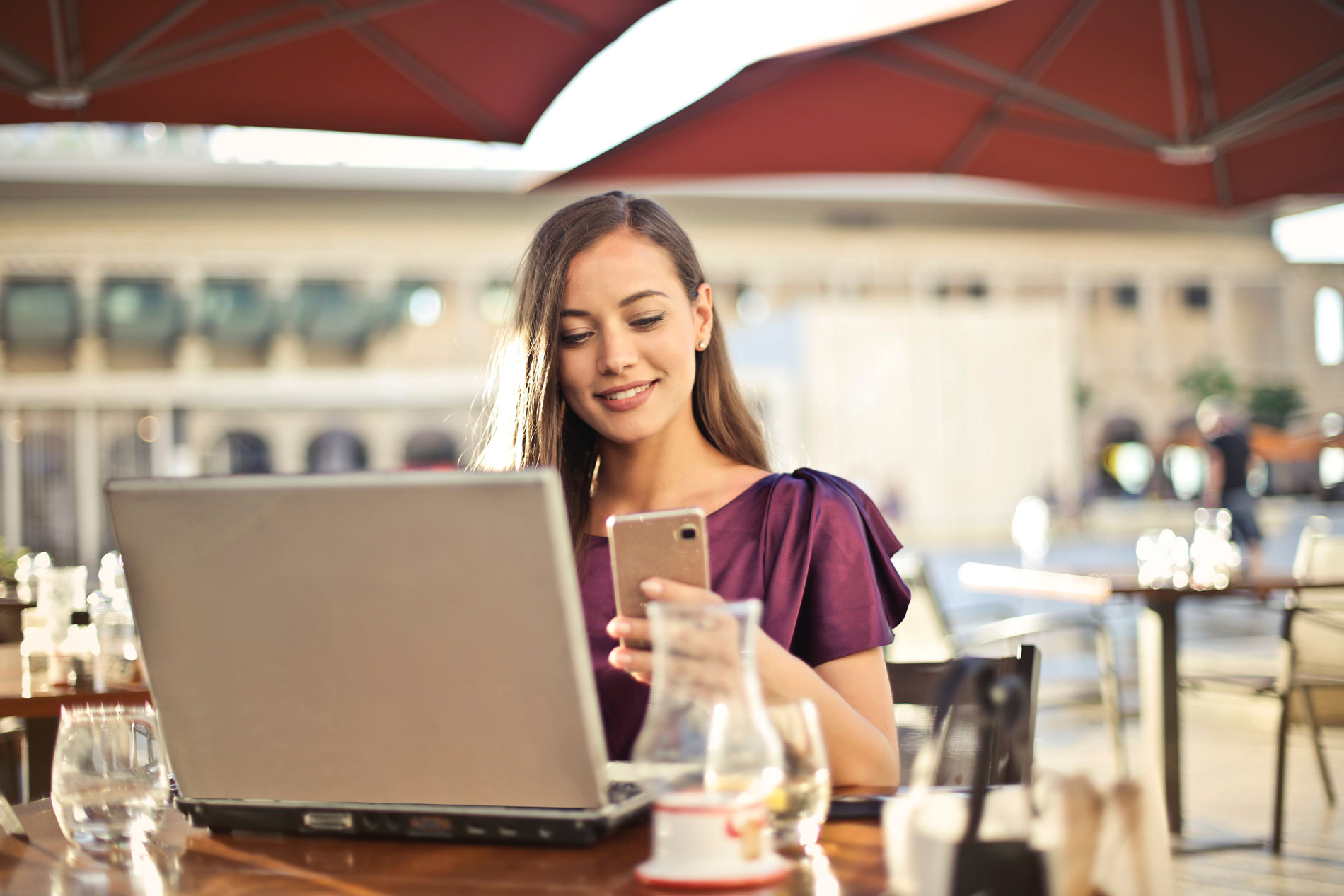 Mujer sonriente revisando su teléfono móvil mientras trabaja en su laptop en una terraza, simbolizando la gestión flexible y accesible de sus seguros y gastos médicos